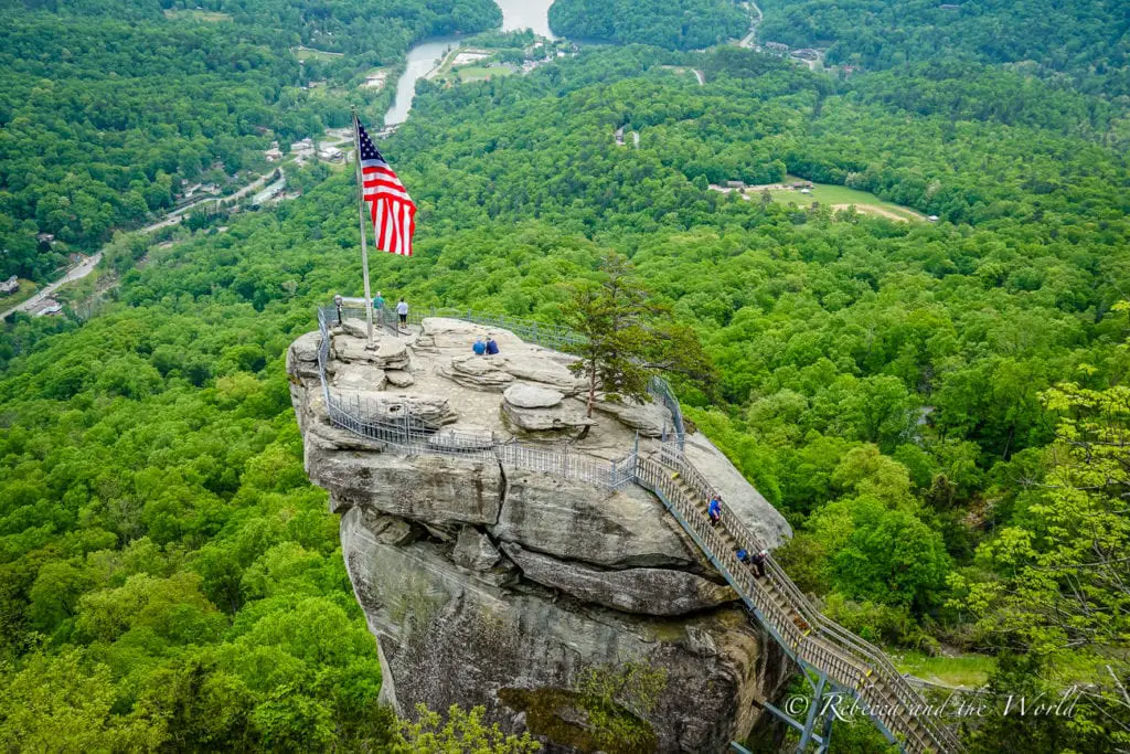 An imposing rock formation with a large American flag at the peak. Visitors can be seen on the lookout points, connected by a narrow walkway, with dense green forest and a winding river visible in the valley below. Asheville is home to plenty of outdoors activities, like Chimney Rock State Park.