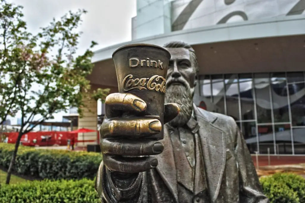 A bronze statue of a man holding a cup with the "Drink Coca-Cola" logo, with a modern building and red tents in the background, reflecting the region's connection to the beverage industry. In Atlanta, one of the most fun things to do is the Coca-Cola museum.