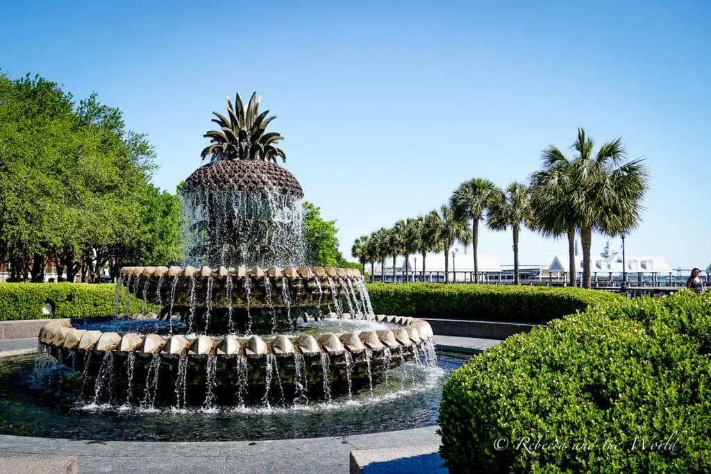 A striking fountain with water cascading down tiers shaped like a pineapple, a common symbol of hospitality, set in a public space with palm trees and a clear blue sky overhead. Charleston is a must-stop city on a USA Deep South road trip