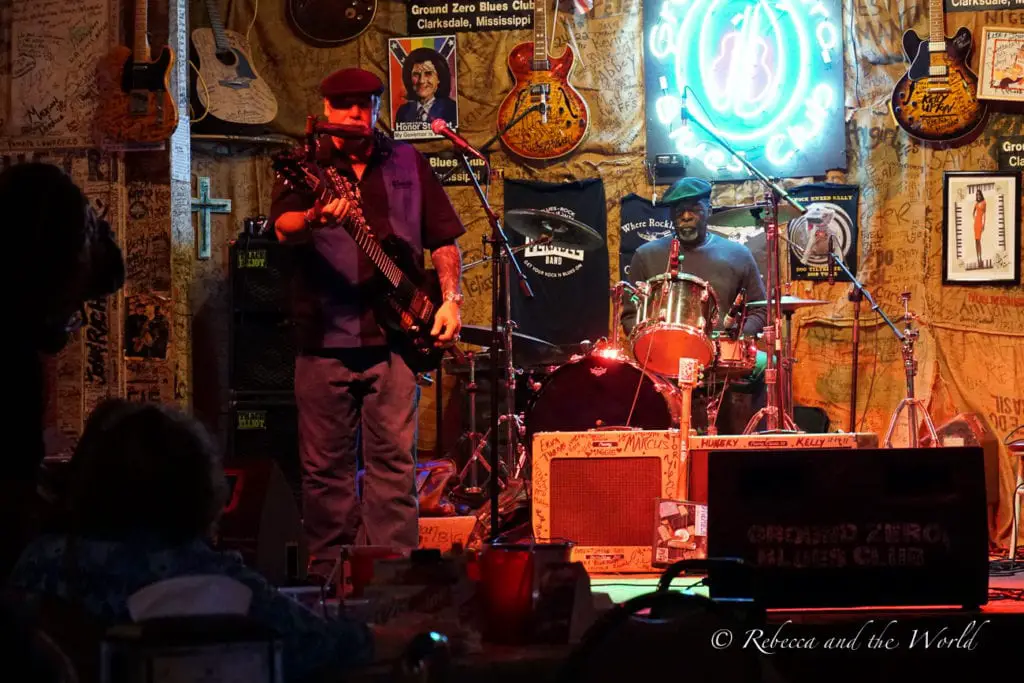 A dimly-lit blues club interior with a guitarist and drummer performing on stage, surrounded by walls covered in music memorabilia and neon signs. Clarksdale in Mississippi is the birthplace of the blues and you'll be able to catch live music most nights of the week.