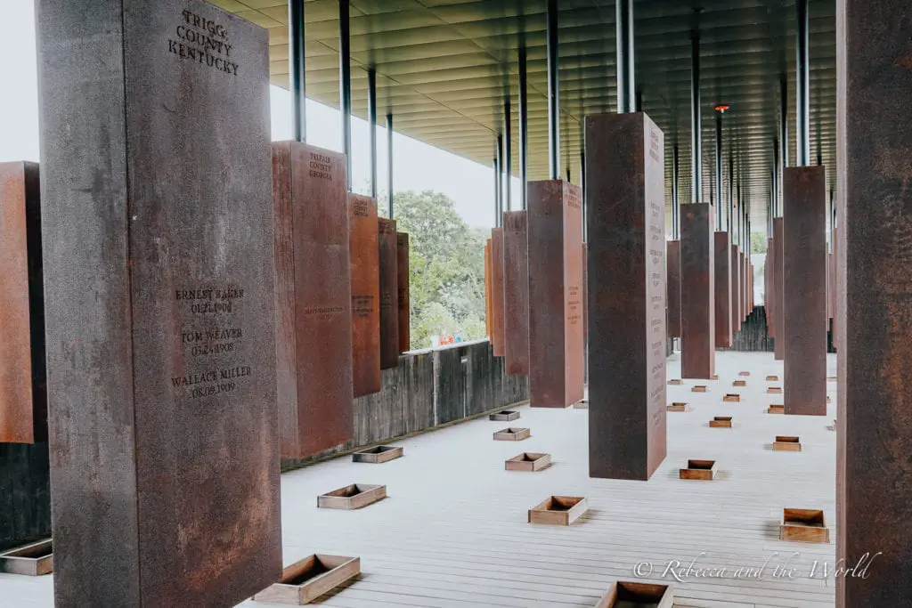A somber memorial space with a series of tall, rust-colored metal columns suspended above the ground, representing the victims of lynching; the names and dates are inscribed on the columns.
