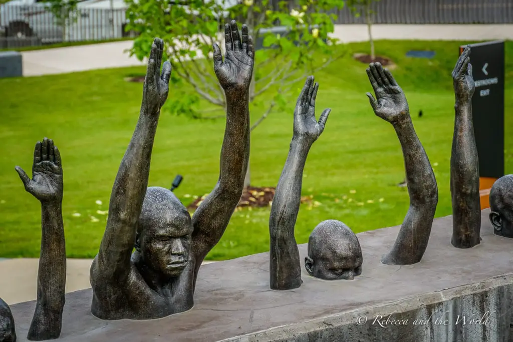 A sculpture depicting raised hands and a head emerging from the ground set against a backdrop of a neatly kept lawn. This is at the harrowing Legacy Museum and National Memorial for Peace and Justice in Montgomery, AL.