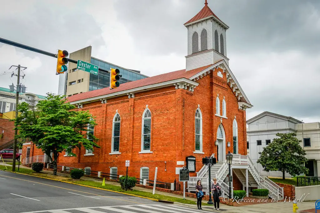 A historic red brick church with white trim, a steeple, and an external staircase. Two individuals are walking by the church on a city street with traffic lights overhead. The Dexter Avenue King Memorial Baptist Church is one of the best places to visit in Montgomery Alabama.