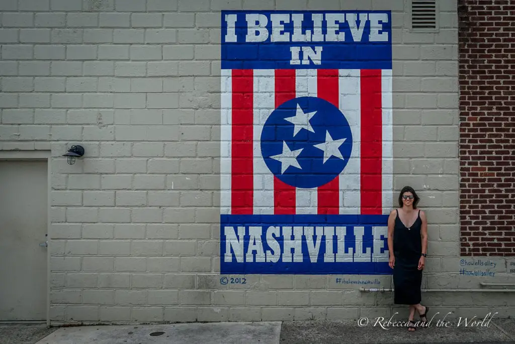 A mural on a brick wall featuring an American flag design with the words "I BELIEVE IN NASHVILLE," with a woman - the author of this article - standing in front of it. This mural is famous in Nashville, TN, and should be on your Deep South road trip itinerary.