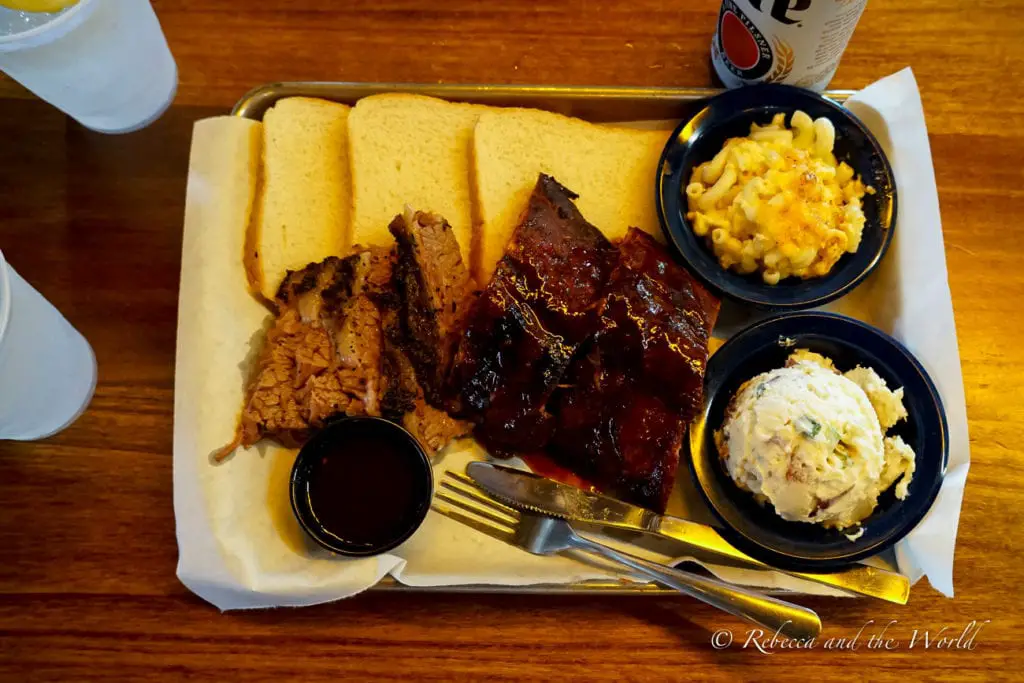 A meal of barbecue consisting of sliced brisket, ribs, pulled pork, slices of white bread, with sides of macaroni and cheese, and potato salad, accompanied by a cold beer in a can, served on a tray.