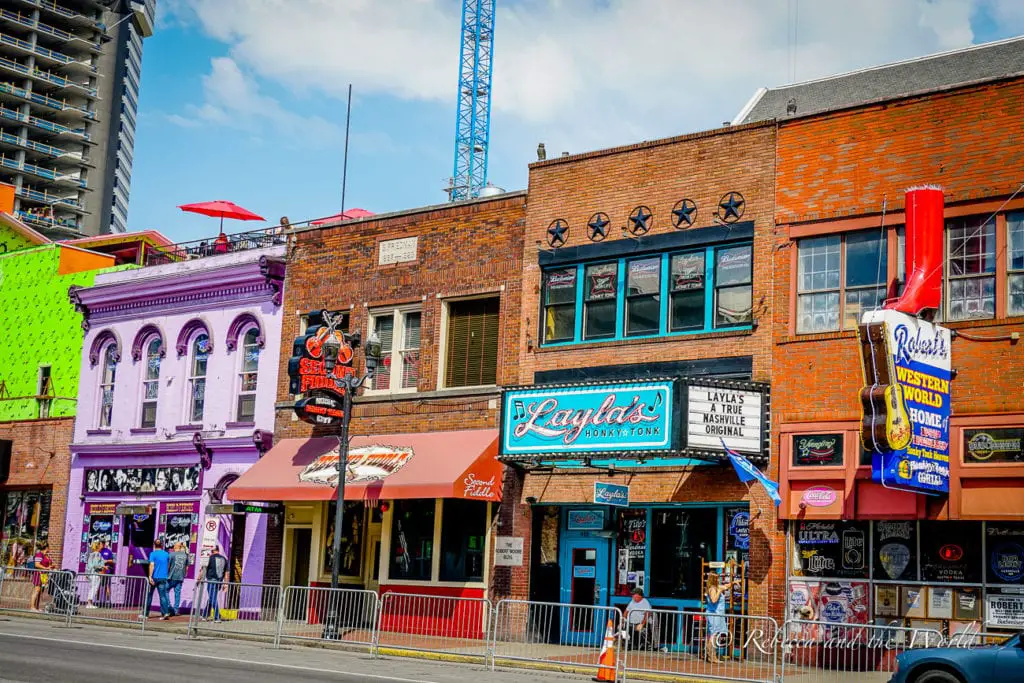 A bustling street in Nashville with historic buildings, neon signs, and various establishments including bars and honky-tonks, reflecting the city's vibrant music scene.