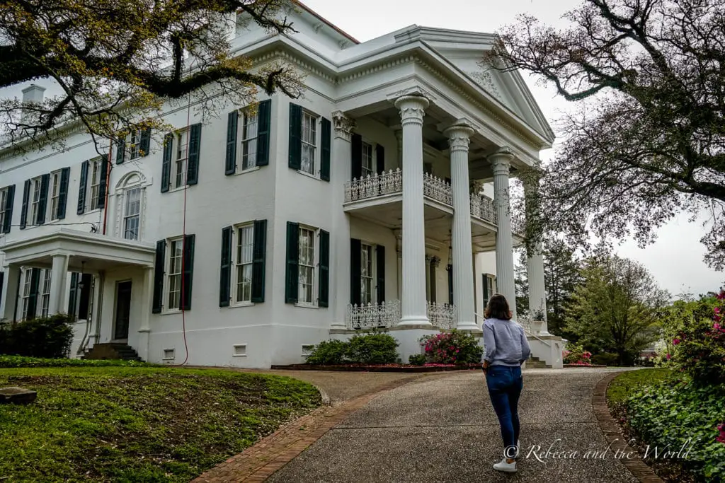 A person - the author of this article - walking towards a stately white mansion with tall columns, black shutters, and a sprawling front porch, set against large trees and a lush green lawn.