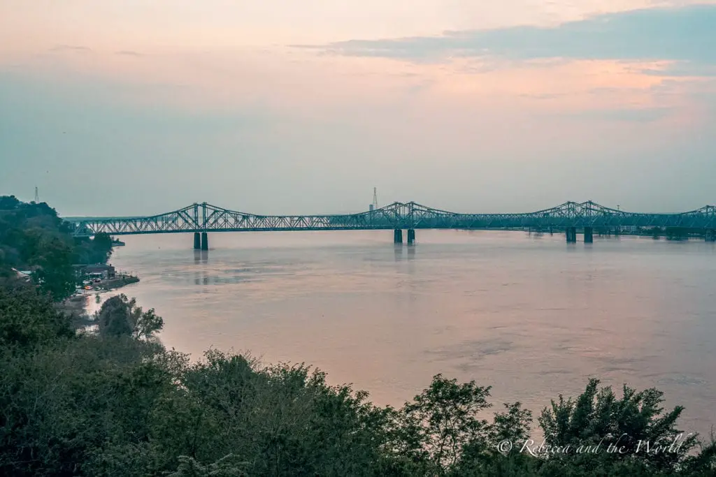 A serene view of a wide river with a long, multi-arched bridge spanning across. The bridge's reflection is visible in the calm water, and trees line the riverbank. The views over the Mississippi River in Natchez are stunning, especially at sunset.