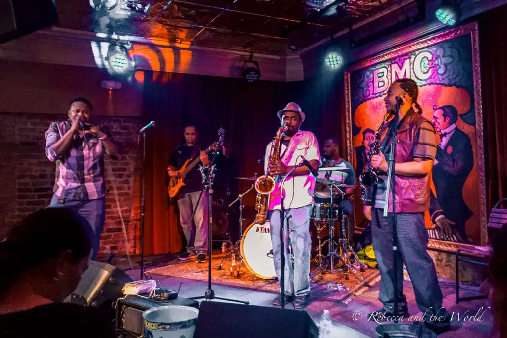 A live music scene inside a dimly lit club with a band performing; a vocalist at the center with a harmonica, a saxophonist, and other band members are visible on stage, with colorful stage lights and the "BMC" logo in the background. Catch plenty of live music on a stop in New Orleans on a Deep South road trip.