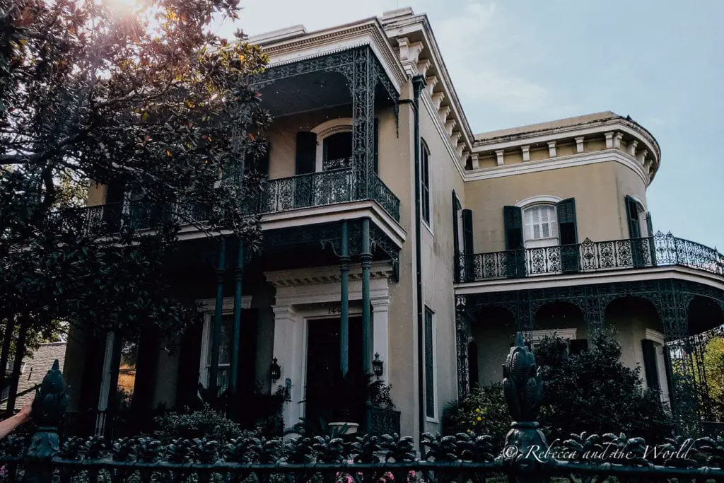 A stately two-story house with classical architecture featuring ornate balconies, tall columns, and a dark facade. Mature trees and a decorative iron fence surround the property. Wandering the Garden District to see the incredible homes is one of the best things to do in New Orleans.