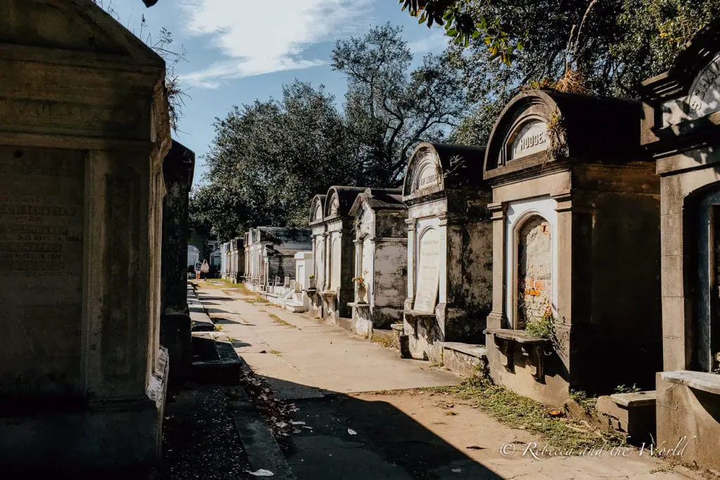 An avenue through an old cemetery with above-ground tombs and vaults, showing the distinctive burial practice of the region. Sunlight filters through the trees, highlighting the historical gravestones. Spook yourself at one of New Orleans' cemeteries.