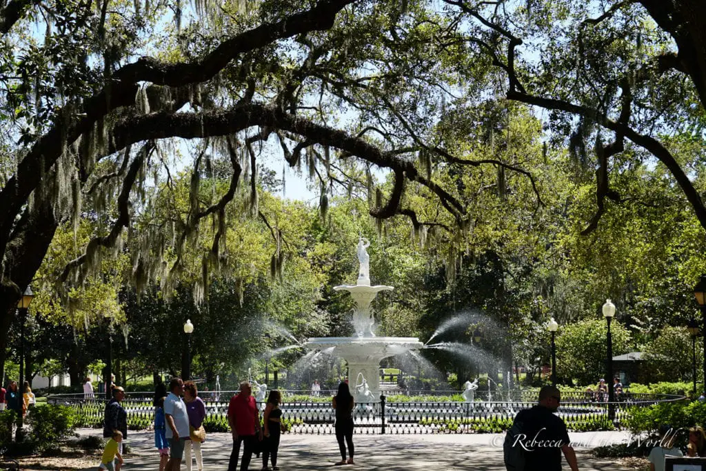 A public park scene with people walking and conversing near a white ornamental fountain spraying water, framed by the overarching branches of live oak trees draped with Spanish moss. Savannah is one of the most beautiful places to visit in the Deep South, with gorgeous gardens, squares and fountains.