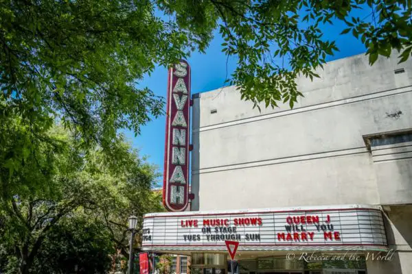 The vintage red and white marquee of the Savannah Theatre with "Live Music Shows" and "Queen J Will You Marry Me" displayed. The theater's name is brightly lit on the vertical sign, nestled among lush green trees. Savannah is a must-visit destination on a Deep South road trip.