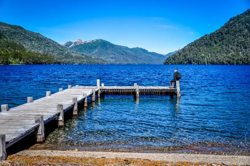 A man in black sits on the end of an L-shaped wooden pier at a blue lake near Bariloche in Argentina. In the background are tree-covered mountains. This lake can be seen on the Ruta de los Siete Lagos (7 Lakes Route) in Argentina.