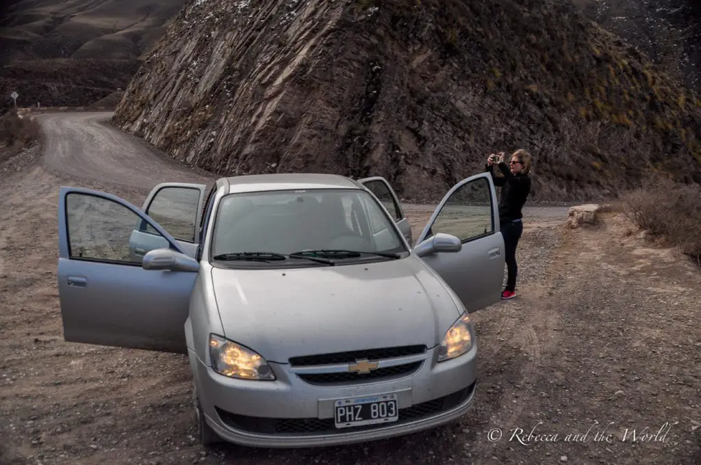 A small grey car is parked on the side of a dirt road, with all its doors open and the headlights on. A woman in black stands by the car taking a photo of an unknown object. Travel insurance for Argentina can sometimes include car insurance for rental cars.