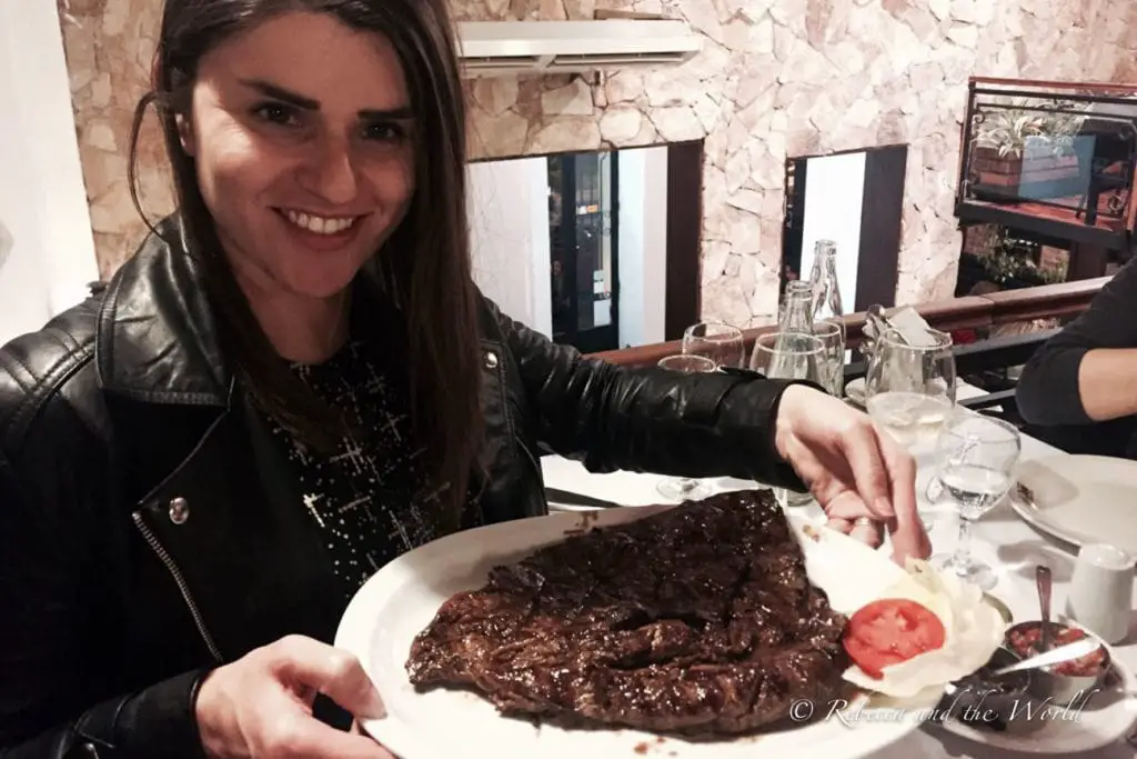 A woman in a black leather jacket smiles at the camera as she holds up a plate with a large piece of cooked steak on it. Food in Argentina is one of the best reasons to visit.