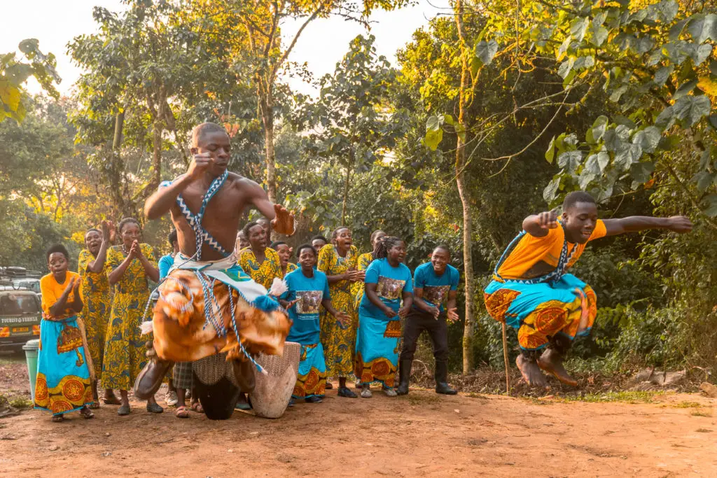 A group of people in vibrant traditional African clothing performing a dance in a clearing surrounded by trees. Two dancers are in mid-motion, with one leaping high off the ground.