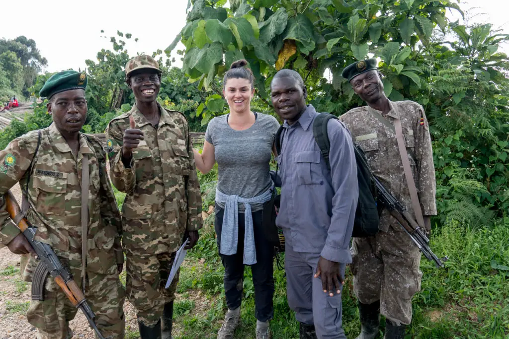 Four individuals in camouflage military uniforms, some carrying rifles, posing for a photo with a smiling woman - the author of this article -  in casual clothing. They are standing in a lush, green area. All gorilla trekking groups are accompanied by armed guides and you can also hire a porter.