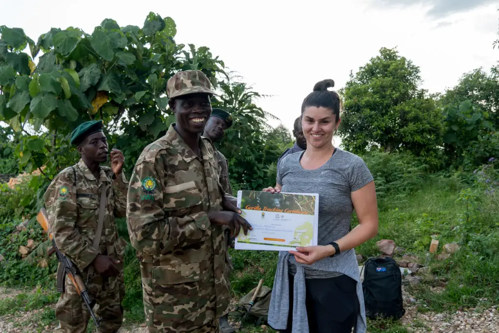 A woman - the author of this article - holding a certificate stands with four men in camouflage uniforms. They are outdoors, with trees and foliage in the background.