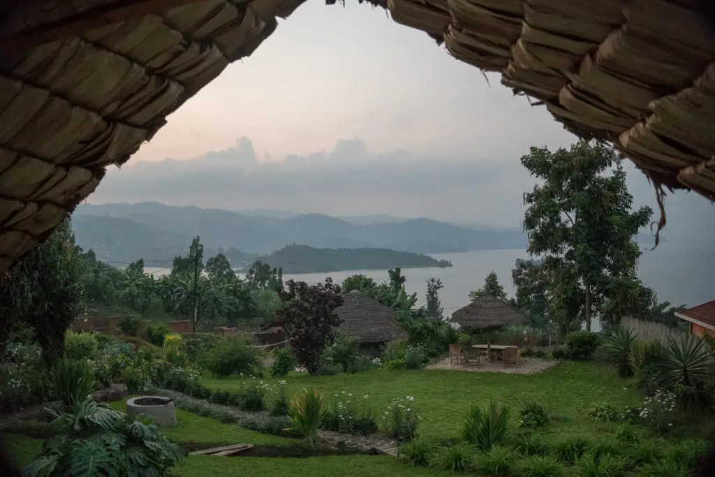 A view from under a thatched roof looking out over a lush garden with a lake in the distance and hills on the horizon, taken at dusk. Gorilla trekking Uganda versus Rwanda? Both have their pros and cons - but whichever country you choose you'll have an amazing experience.