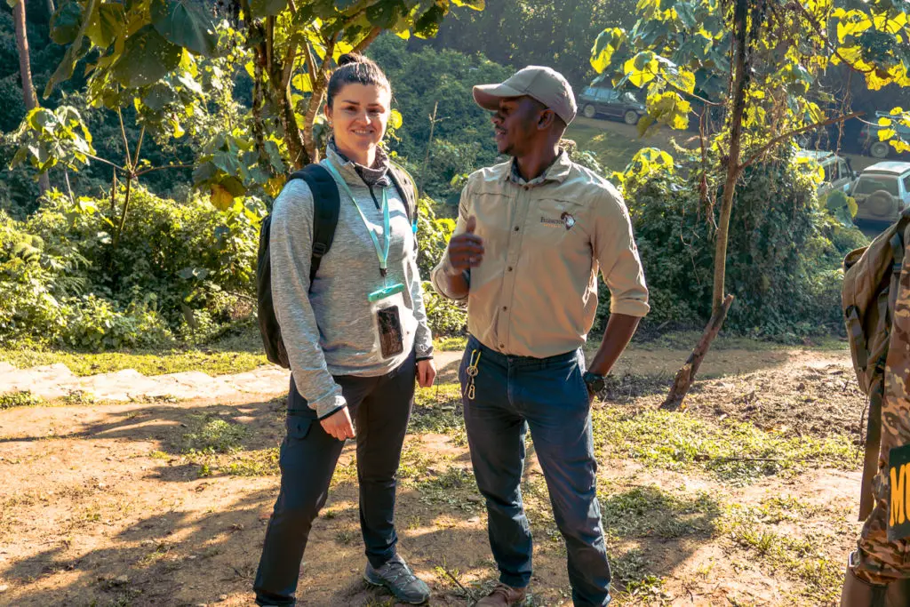 A woman - the author of this article - in casual hiking attire with a backpack is standing next to a man in a khaki shirt and cap, who appears to be a guide. They are conversing and surrounded by lush vegetation. Wondering what to wear gorilla trekking? This gorilla tracking packing list will help.