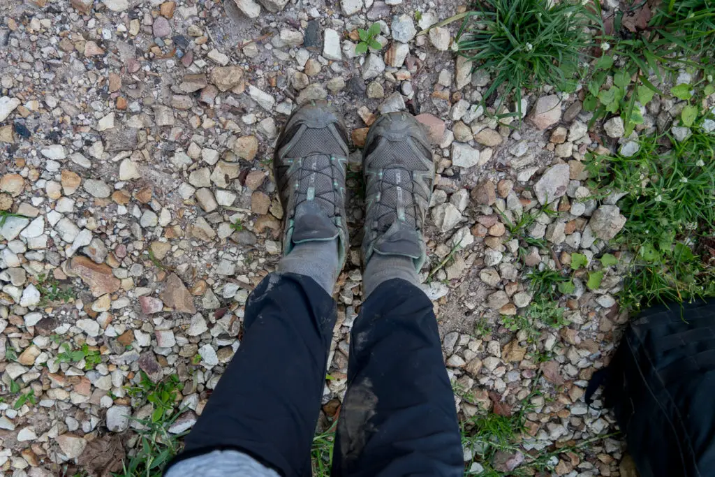 An image from above of a person's lower legs and feet are shown, wearing muddy hiking shoes and black pants. The feet are standing on a gravel path with scattered green grass to the side. The socks are tucked over the person's pants. Wondering what to wear when you first meet gorillas in the wild? This gorilla trekking packing list will help. Tip: tuck your pants into your socks to keep bugs and mud out!