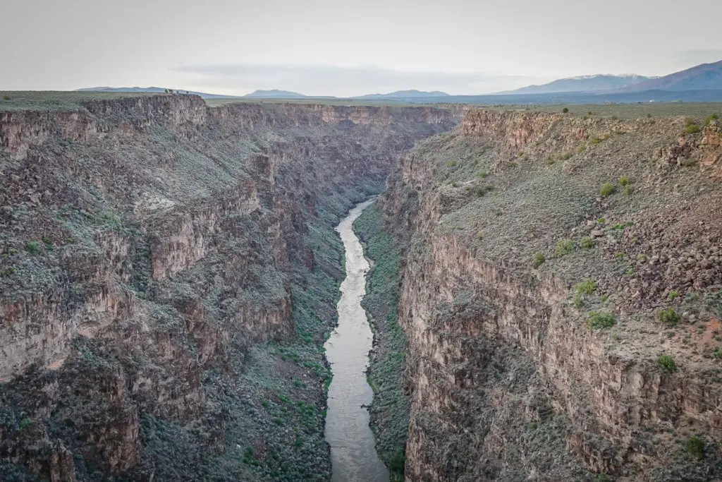 A deep canyon - Taos Gorge in New Mexico - with a narrow river running through it, viewed from a high vantage point. The canyon walls are steep and rocky, with sparse vegetation, and the sky is dusky, suggesting either dawn or dusk.