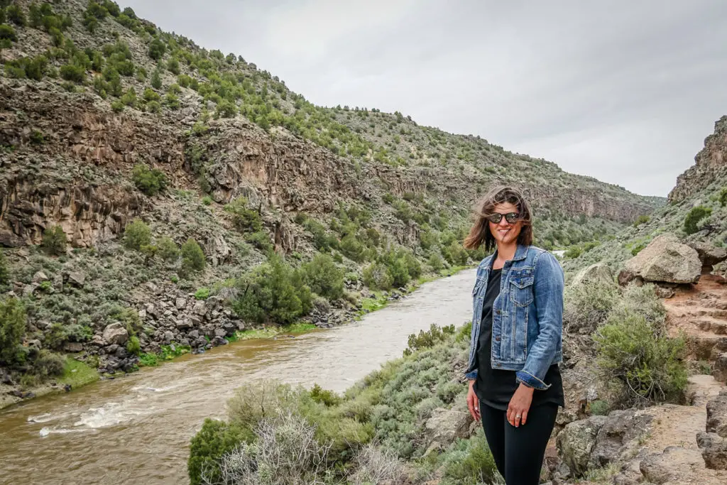 A smiling woman - the author of this article - with sunglasses and a denim jacket standing on a rocky overlook. She is facing the camera with a river and rugged canyon walls in the background. This photo was taken at Taos Canyon in New Mexico.