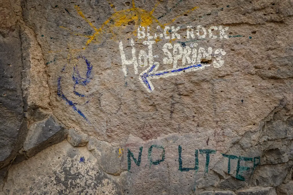 A close-up of a rock wall with graffiti indicating directions to "Black Rock Hot Springs" with an arrow. Below, another message reads "NO LITTER" in green paint. One of the best things to do in Taos, New Mexico, is soak in hot springs.