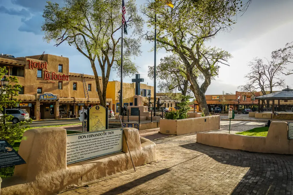 A historic plaza with a large tree in the foreground, adobe buildings, and a clear sky. Signs and flags are visible, and the place appears calm with no visible people. Taos should definitely be on your New Mexico road trip itinerary.