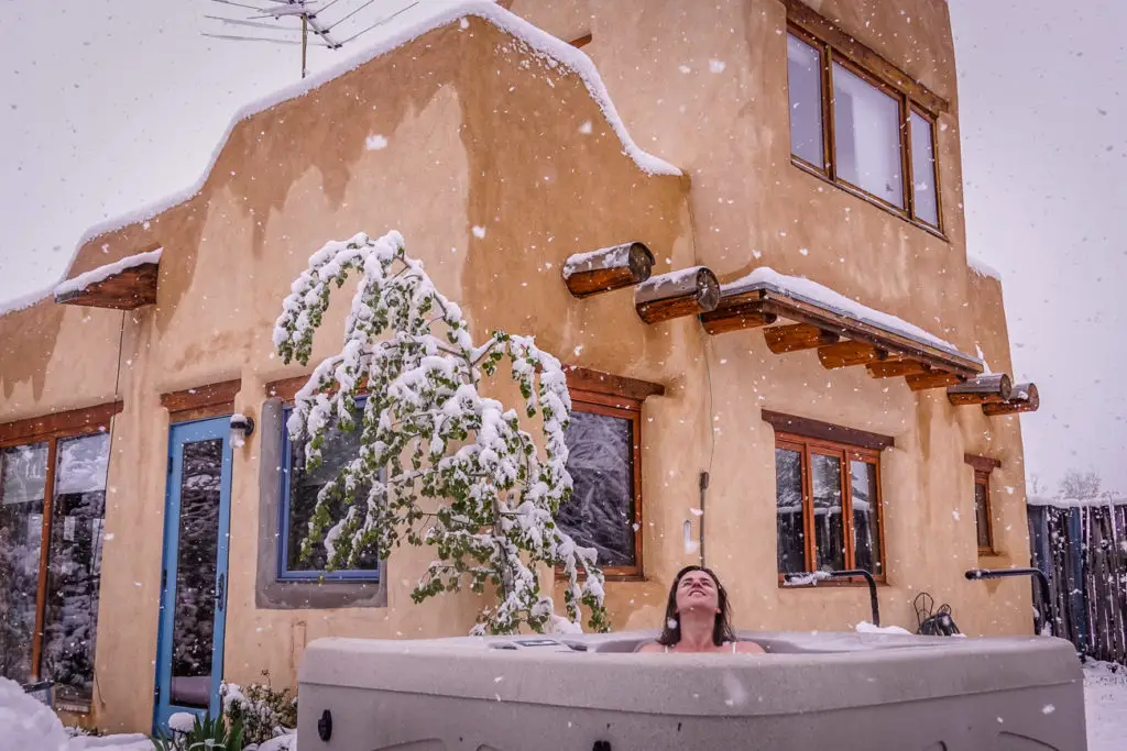 A person relaxes in an outdoor hot tub, surrounded by snow-covered ground and an adobe-style house, in a serene New Mexico winter setting.