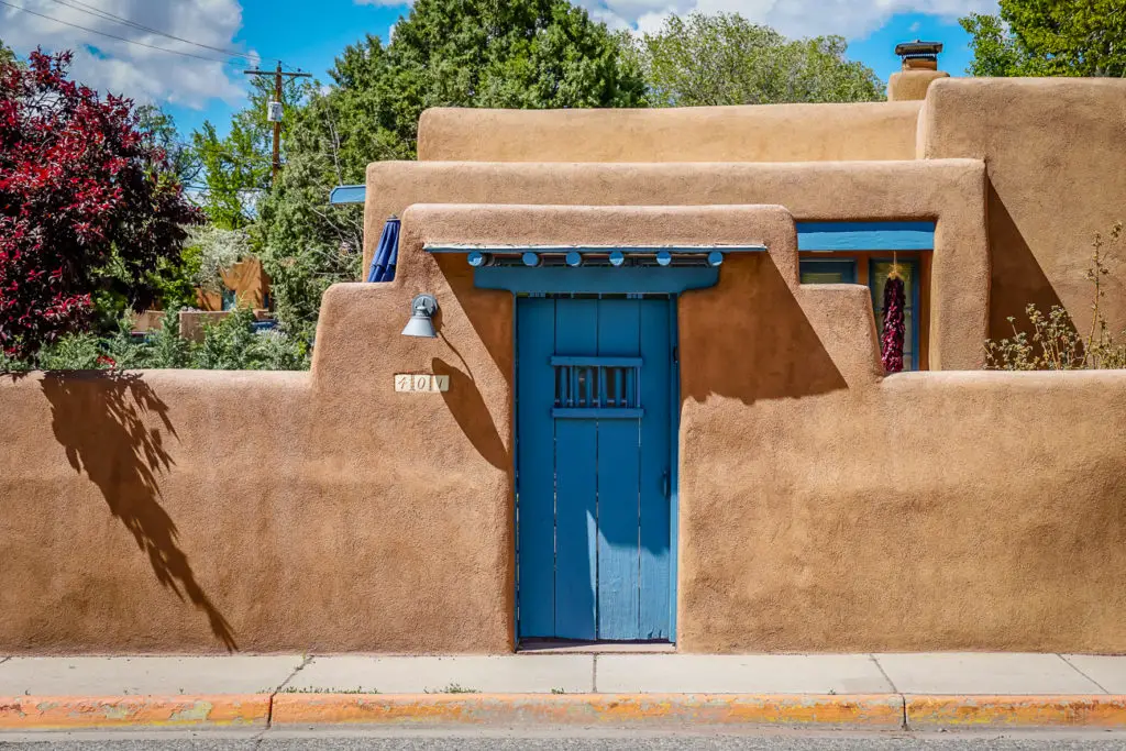 A charming adobe building in Santa Fe, New Mexico, with a bright blue door and matching trim. The house is surrounded by a tall adobe wall, and there are lush plants and a tree casting a shadow on the facade.