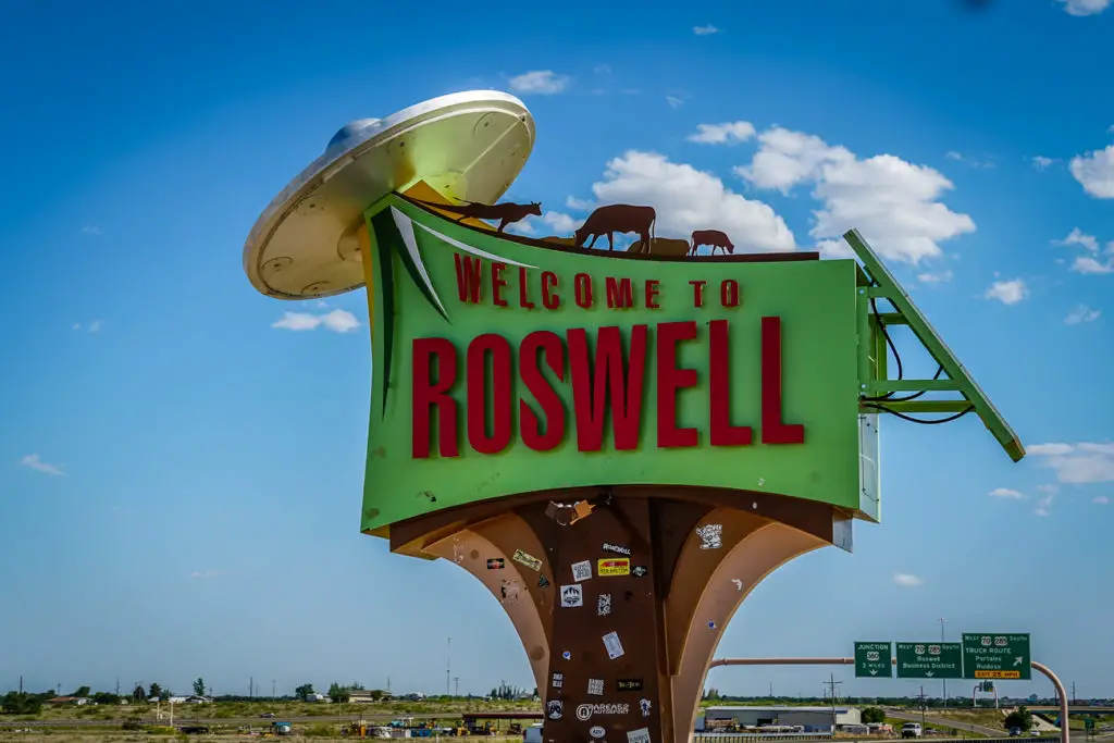 The iconic "Welcome to Roswell" sign featuring a spaceship and silhouettes of cows, against a clear blue sky with directional signs in the background. Roswell is one of the quirkiest places to visit in New Mexico!