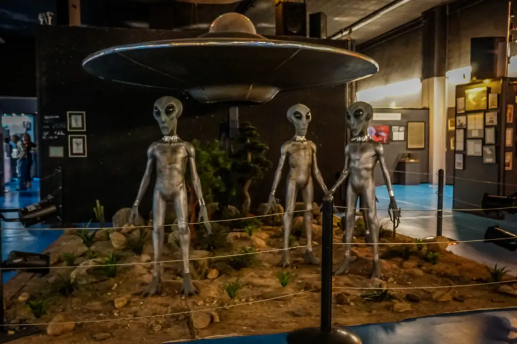 A dimly lit exhibit featuring three life-sized gray alien figures standing in front of a mock-up of a silver flying saucer within an indoor setting. This is the International UFO Museum and Research Center in Roswell, New Mexico - a quirky place to add to your New Mexico itinerary!