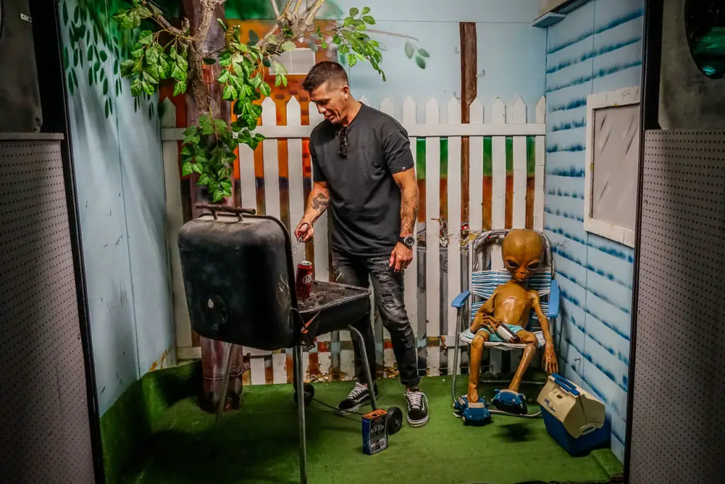 A man standing by a grill with a brown alien figure seated nearby in a casual backyard setting, creating a humorous tableau of an alien barbecue. This is Alien Zone in Roswell, a must-visit in New Mexico if you're interested in UFOs!