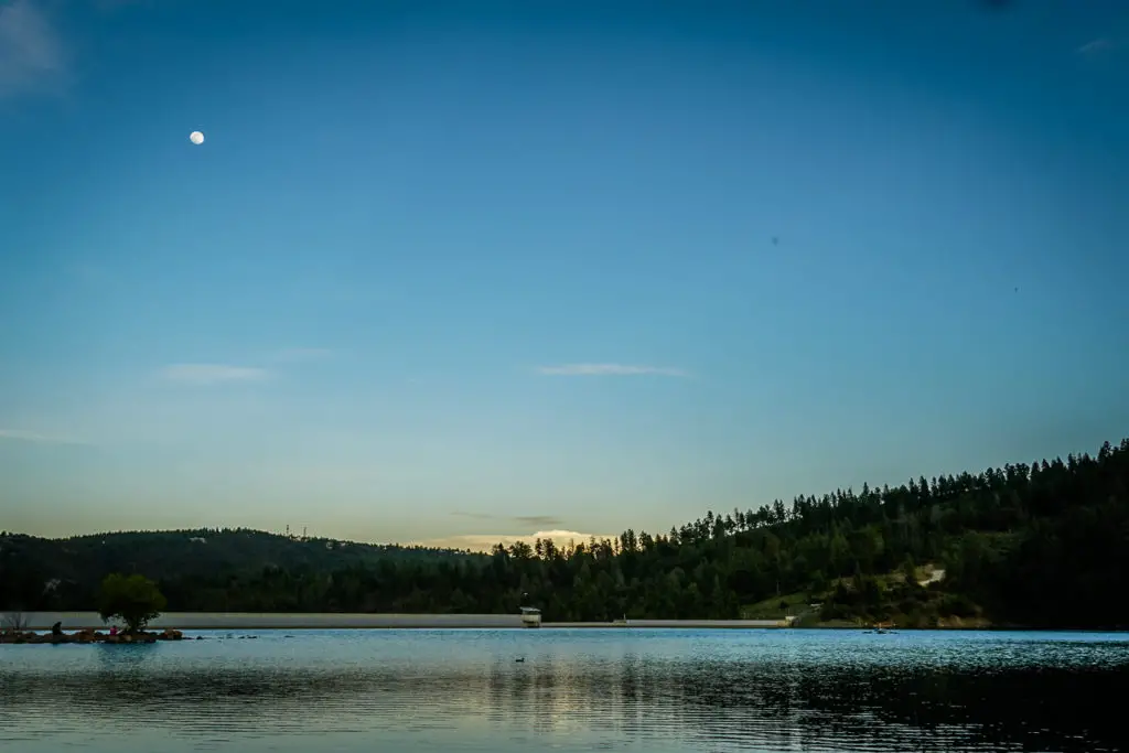 A serene lake with the moon visible in the twilight sky, surrounded by a forested landscape reflecting in the calm water. This is Grindstone Lake in Ruidoso, New Mexico.