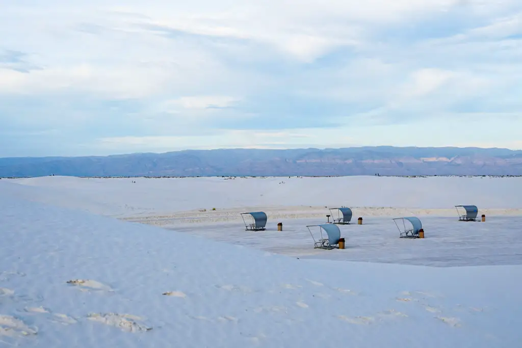 Picnic shelters partially buried in white sand dunes, with a mountain range in the distance and overcast sky above, in White Sands National Park, New Mexico.