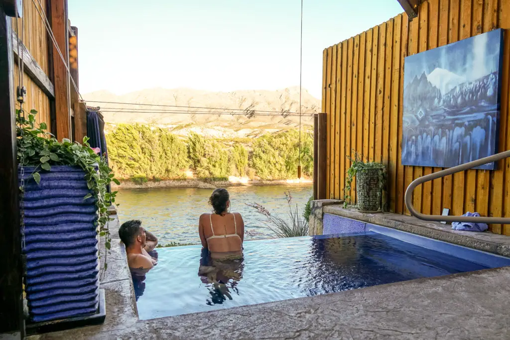 Two individuals relaxing in an outdoor hot tub in Truth or Consequences, New Mexico, with a scenic view of mountains in the background, partially obscured by wooden fencing and foliage.