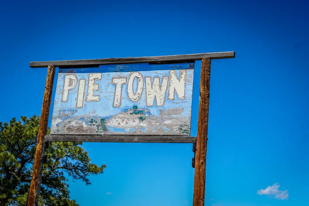 A weathered wooden sign that reads "Pie Town," indicating the historic community in New Mexico known for its pies, set against a bright blue sky.