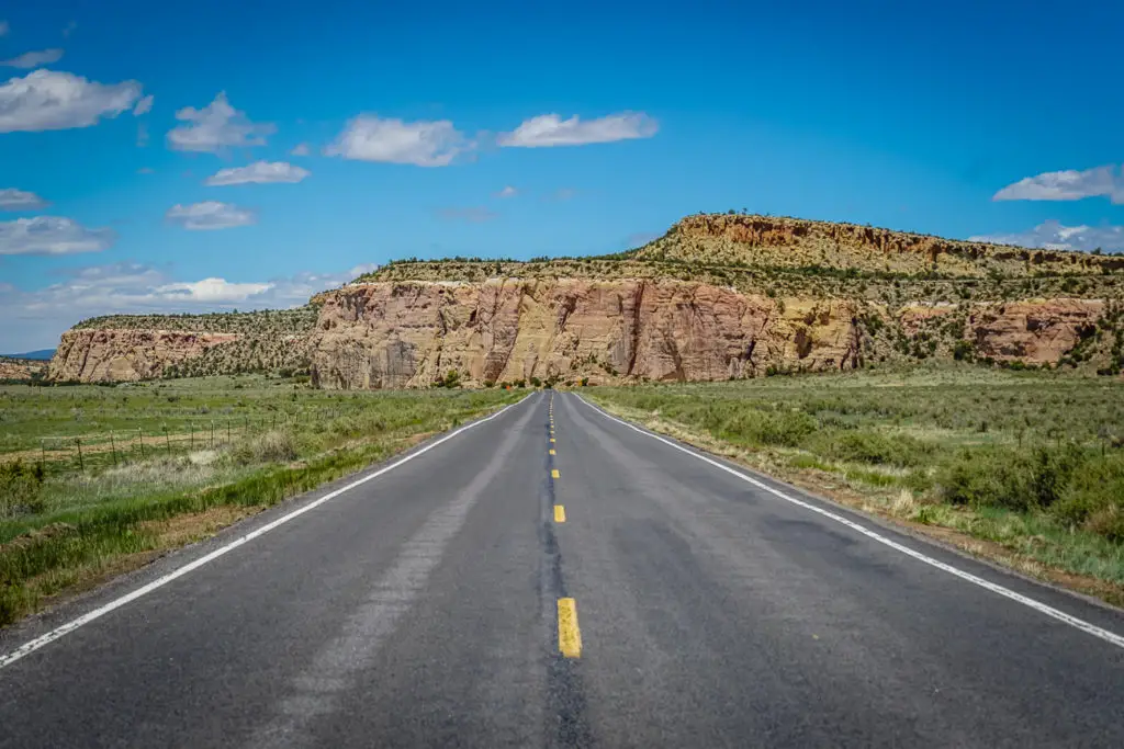 A straight two-lane highway leading towards large rock formations under a bright blue sky dotted with fluffy white clouds. The landscape on either side of the road is covered with sparse green shrubbery. A New Mexico road trip is a great way to see this state.