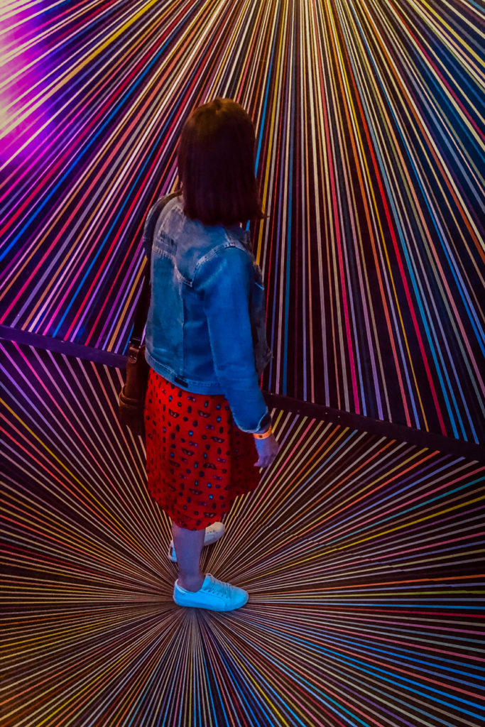 A person - the author of this article - viewed from behind, standing in front of a vibrant, multicoloured light installation that radiates from a central point. The individual is wearing a denim jacket and a red dress with a black pattern, standing in a dark room illuminated by the light exhibit. This is Meow Wolf, one of the best things to do in Santa Fe, New Mexico.