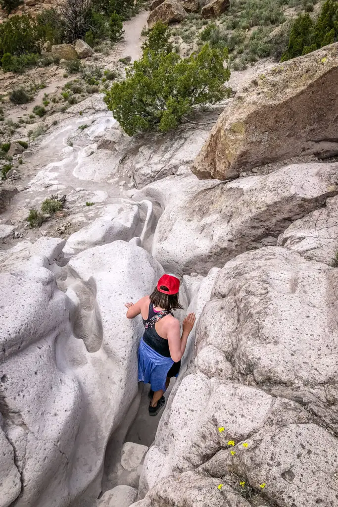 A woman - the author of this article - with a red cap and hiking pants climbing down a narrow, carved path in the white rocky terrain with sparse vegetation around. This is Tsankawi, just outside of Santa Fe, New Mexico.