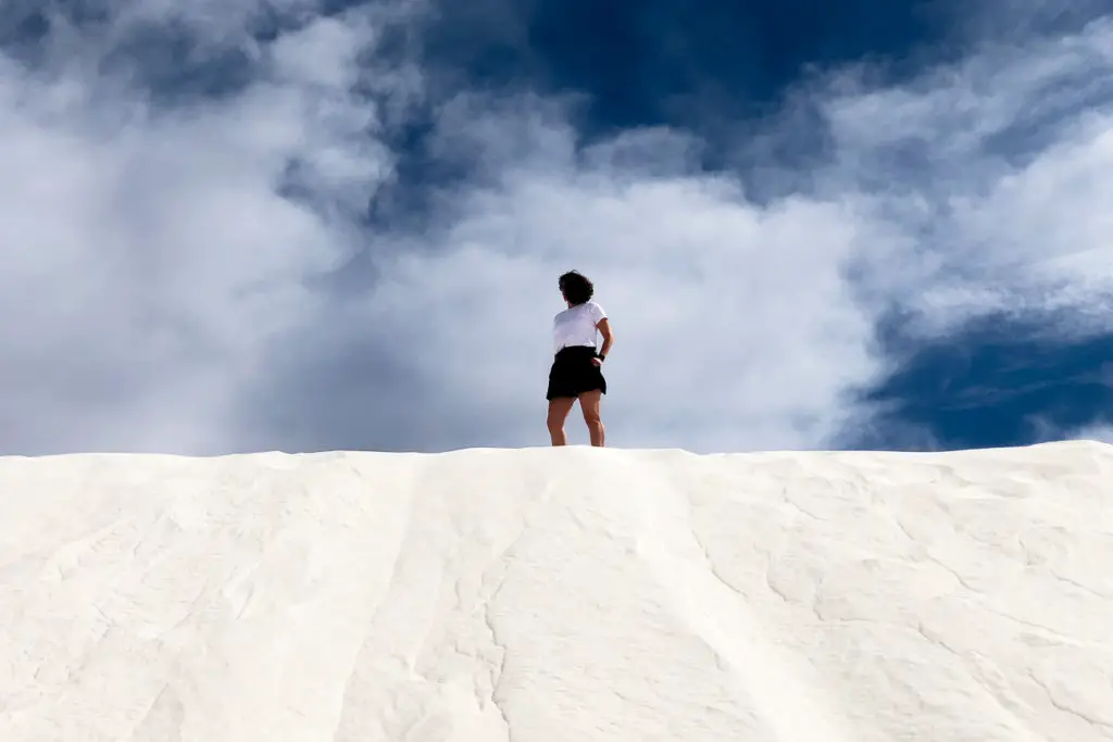 A person - the author of this article - standing on the crest of a white sand dune, looking into the distance with a dynamic sky of white clouds and blue sky above. This is White Sands National Park, a place that should definitely be on any New Mexico road trip itinerary.