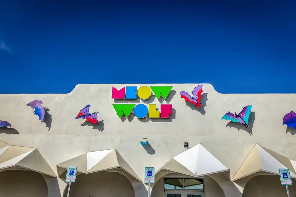 The colorful and stylized "Meow Wolf" sign on the facade of a building, with abstract figures resembling bats in flight above the lettering. Meow Wolf is one of the coolest places to visit in Santa Fe, New Mexico.