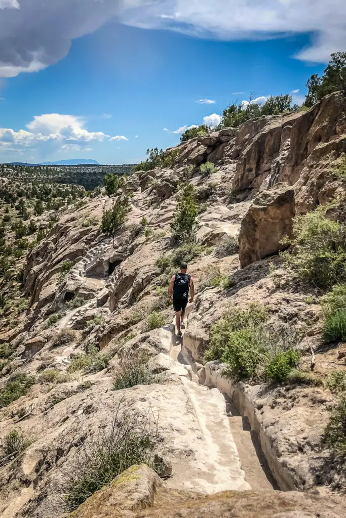 A person - the author's husband - with a backpack walking along a narrow trail carved into a rocky landscape with bushes and a clear sky above. This is Tsankawi, just outside of Santa Fe, New Mexico.