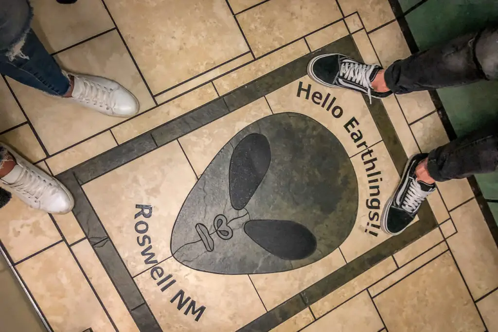 A floor inset with a graphic saying "Hello Earthlings" with a cartoonish alien face, located in Roswell, NM, as indicated by the text on the floor, with two pairs of feet standing on either side of the greeting.