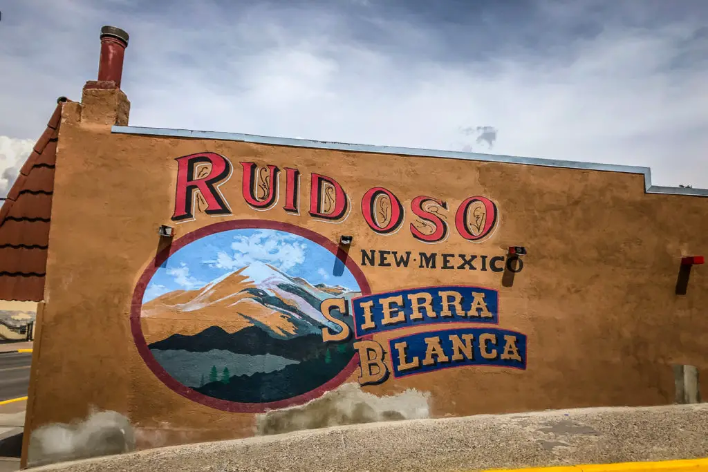 A mural on a building with "RUIDOSO New Mexico Sierra Blanca" depicting a mountain landscape within a large circle, and the rest of the wall in adobe style.