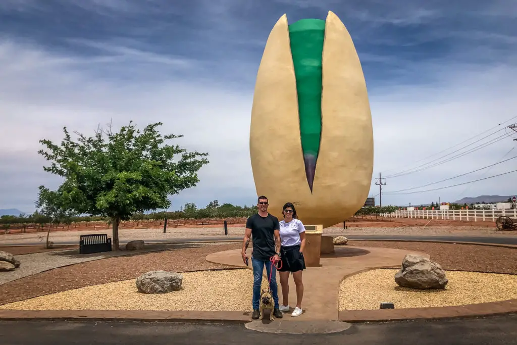 Two people and a dog - the author, her husband and her dog - posing in front of a large sculptural representation of a pistachio at Pistachio Land in New Mexico.