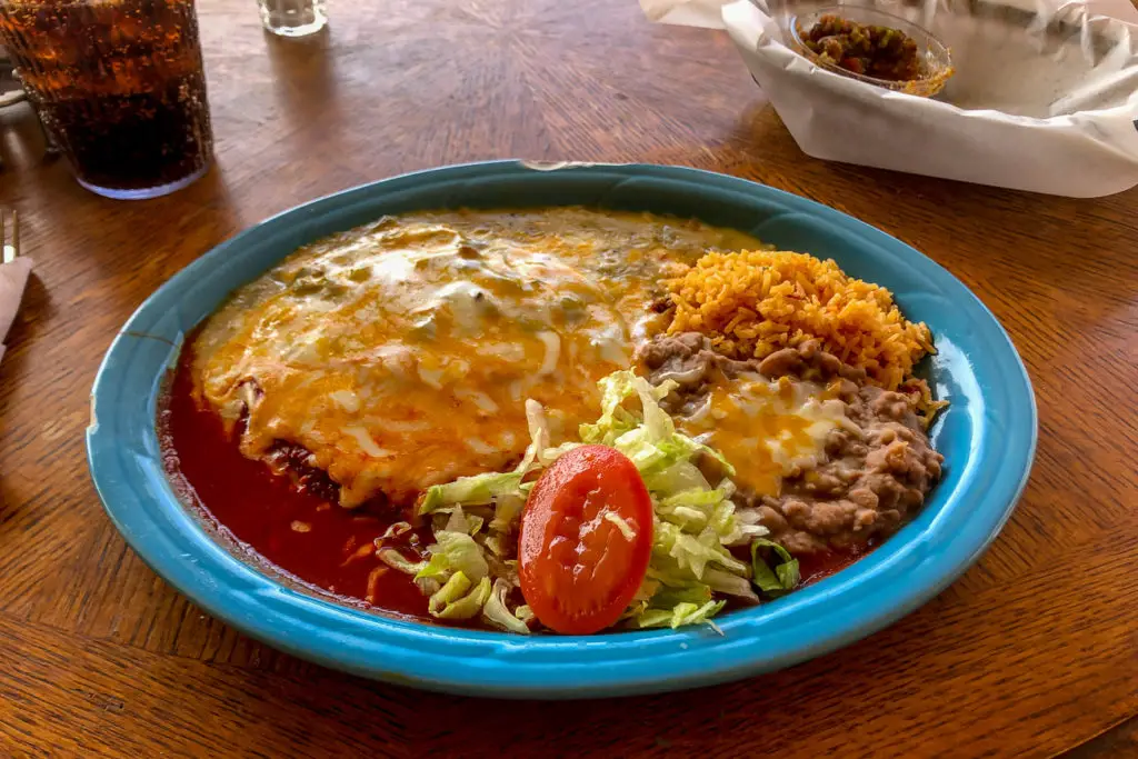 A plate of traditional New Mexican cuisine with an enchilada smothered in cheese and red sauce, served with sides of rice and refried beans, topped with a slice of tomato and shredded lettuce.