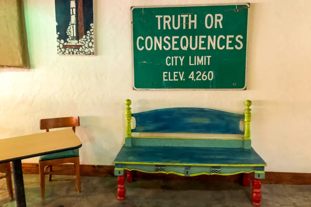 A whimsical indoor scene with a "TRUTH OR CONSEQUENCES CITY LIMIT" sign, with an elevation detail, placed above a brightly painted wooden bench and a small table with a chair. Don't miss the small town of Truth or Consequences on your New Mexico road trip.